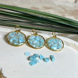 Runder Anhänger „Golden Atlantis Dream“ mit kleinen Larimar-Edelsteinchen in klarem Resin, gefasst in einer 24K vergoldeten Zinklegierung in Shiny Gold. Die Larimarsteine zeigen sanfte Blau- und Weißtöne wie karibische Wellen. Durchmesser ca. 22 mm, handgefertigtes Unikat, fotografiert mit Larimarsteinchen, Muscheln und einer 2-Euro-Münze zur Größenorientierung auf maritimem Untergrund.