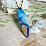 Handgefertigter Surfbrett-Anhänger „Ocean Glide“ aus Maserholz und Epoxidharz in verschiedenen Blau-, und Weißtönen. Der ca. 6,5 × 2 cm große Anhänger wird an einem verstellbaren Lederband (26–46 cm) mit Holzperlen geliefert. Ein maritimes Unikat mit natürlicher Holzmaserung, das die Schönheit des Meeres einfängt.