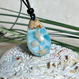 Handgefertigter Tropfenanhänger aus Maserholz und klarem Epoxidharz in Aquatönen. Im Harz sind echte Muscheln und ein kleiner Seestern eingebettet. Durch die besondere Mehrschicht-Gießtechnik wirkt es, als würden die Muscheln im Wasser schweben. Größe ca. 3,8 × 2 cm. Verstellbares Lederband (24–44 cm) mit Holzperlen. Maritimes Unikat.