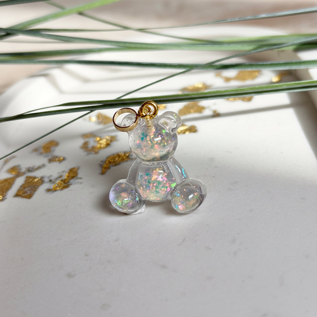 Kleiner Bärchen-Anhänger aus transparentem Harz mit irisierenden Flakes in Regenbogen-Schimmer, goldfarbene Anhängerschlaufe aus Edelstahl. Größe ca. 2,2 × 2,1 cm. Handgefertigtes Unikat; Flakes-Anordnung variiert, vereinzelt kleine Luftbläschen möglich.