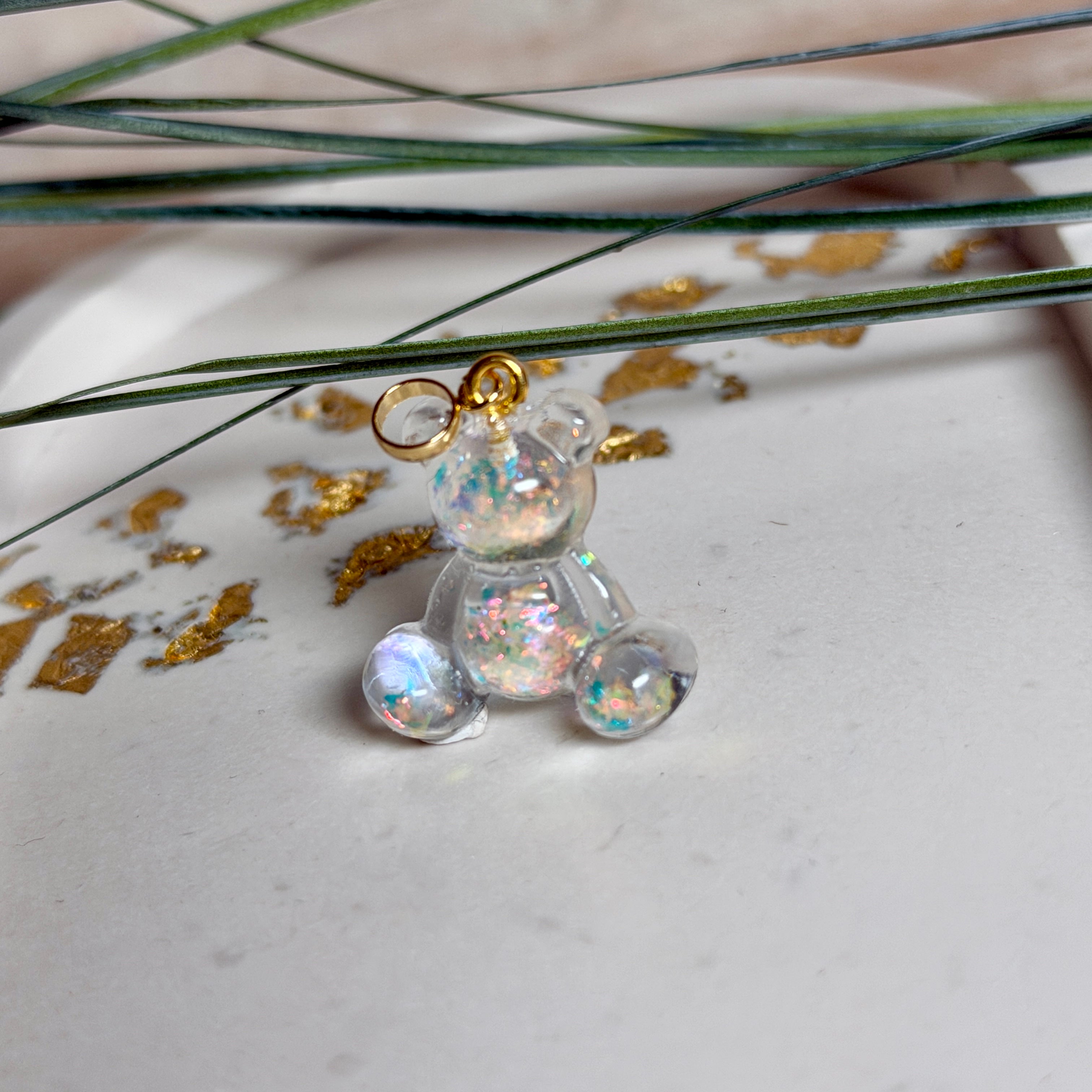 Kleiner Bärchen-Anhänger aus transparentem Harz mit irisierenden Flakes in Regenbogen-Schimmer, goldfarbene Anhängerschlaufe aus Edelstahl. Größe ca. 2,2 × 2,1 cm. Handgefertigtes Unikat; Flakes-Anordnung variiert, vereinzelt kleine Luftbläschen möglich.
