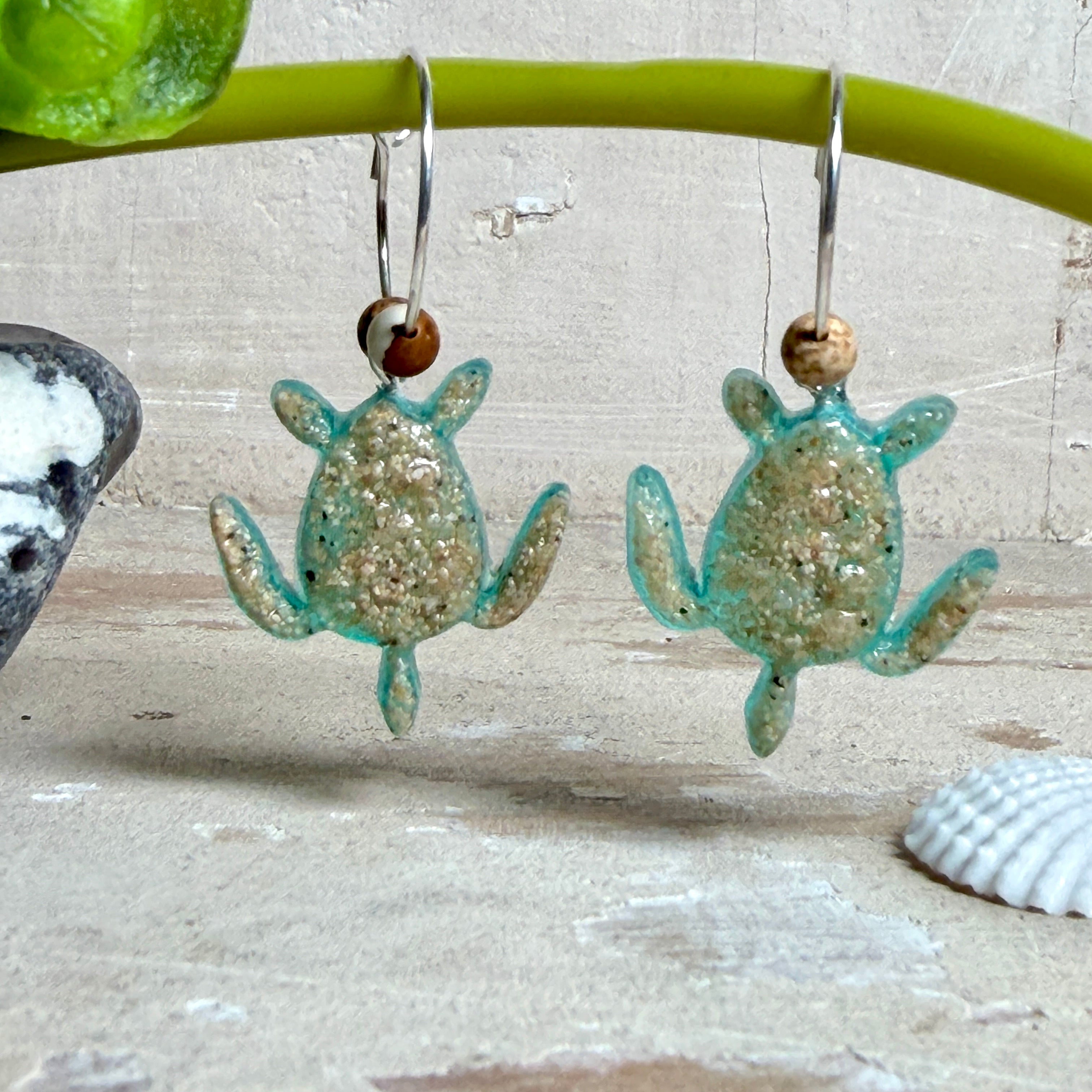 Türkisfarbene Schildkröten-Creolen an feinen 16-mm-Ringen aus 925 Silber, jeweils mit echtem Strandsand im Harz eingebettet und mit kleinen Jaspis-Edelsteinperlen verziert; handgefertigtes Unikat mit sommerlich-maritimem Look, fotografiert als Freisteller, Nahaufnahme oder auf dekorativem Untergrund.