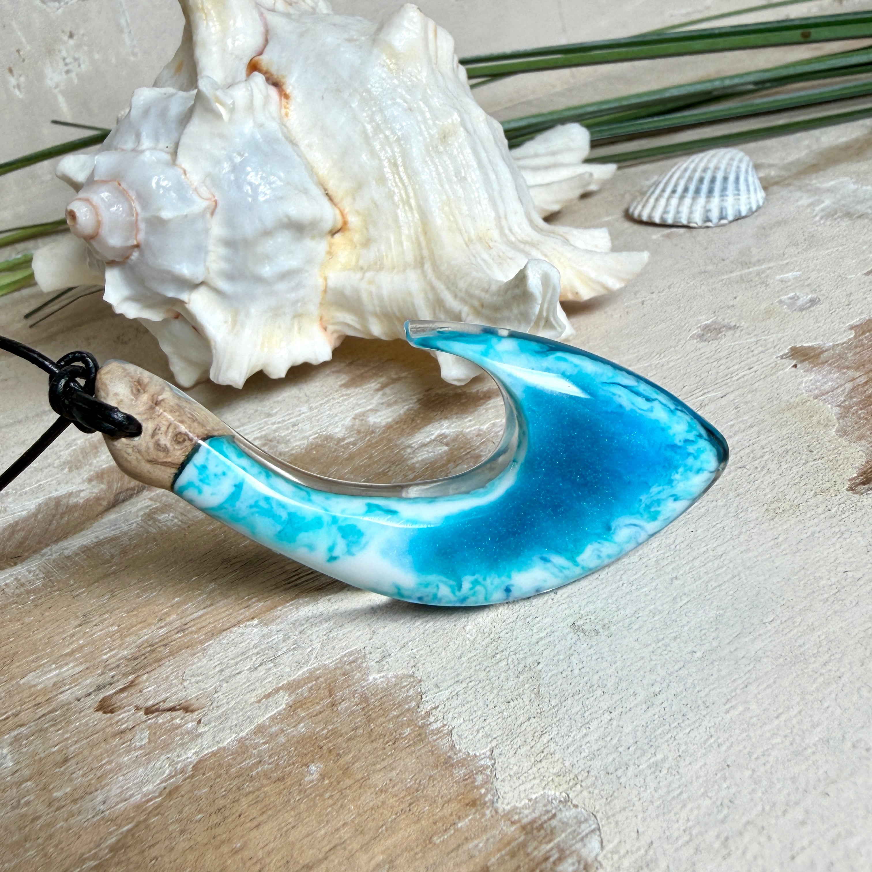 Handgefertigter Haken-Anhänger im Hei-Matau-Stil aus Maserholz und klarem Harz mit Blau-, Türkis- und Weißverlauf, hochglanzpoliert. Oben sichtbares Holz im Harzbereich, getragen an verstellbarem schwarzem Lederband mit Schiebeknoten und Zierperlen. Anhänger ca. 7 × 3 cm, fotografiert mit Muscheln im Hintergrund.