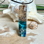 Rechteckiger Stabanhänger aus Maserholz und glasklarem Epoxidharz in tiefen Blau- und Türkistönen mit organischer Lagunen/Riff-Optik. Hochglanzpoliert, ca. 5,3 × 1,2 cm. Der Anhänger hängt an einer schwarzen Lederkette mit Edelstahl-Karabiner und 5-cm-Verlängerung (45 + 5 cm); die Anhängerschlaufe ist ebenfalls aus Edelstahl. Handgefertigtes Unikat, Lieferung in edler Schmuckbox – maritimer Look.