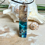 Rechteckiger Stabanhänger aus Maserholz und glasklarem Epoxidharz in tiefen Blau- und Türkistönen mit organischer Lagunen/Riff-Optik. Hochglanzpoliert, ca. 5,3 × 1,2 cm. Der Anhänger hängt an einer schwarzen Lederkette mit Edelstahl-Karabiner und 5-cm-Verlängerung (45 + 5 cm); die Anhängerschlaufe ist ebenfalls aus Edelstahl. Handgefertigtes Unikat, Lieferung in edler Schmuckbox – maritimer Look.