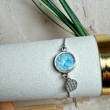 Verstellbares Edelstahlarmband mit Zirkoniasteinchen und Schiebeverschluss und rundem Anhänger (ca. 2,5 × 1,7 cm) mit echten Larimar-Edelstein-Splittern in Harz. Zusätzlich ein Charm „Blume des Lebens“ aus 925 Silber. Handgefertigtes Unikat in Türkis- und Meeresblautönen – Schmuckstück in Nahaufnahme bzw. als Tragebild am Handgelenk.