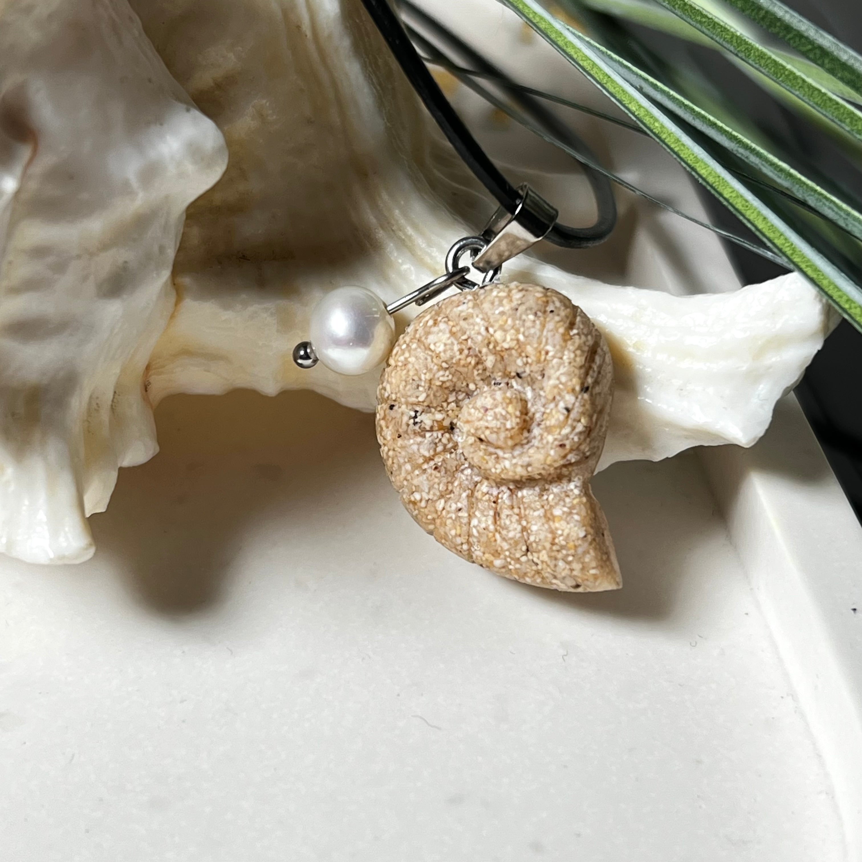 Handgefertigter Ammonit-Anhänger aus echtem Meersand & Harz mit filigraner Edelstahlöse und einer edlen Süßwasserperle. Maritimer Schmuck, inspiriert von der Kraft und Schönheit des Ozeans. Jedes Stück ein Unikat. Präsentiert auf einer Muschel, mit Lederkette.