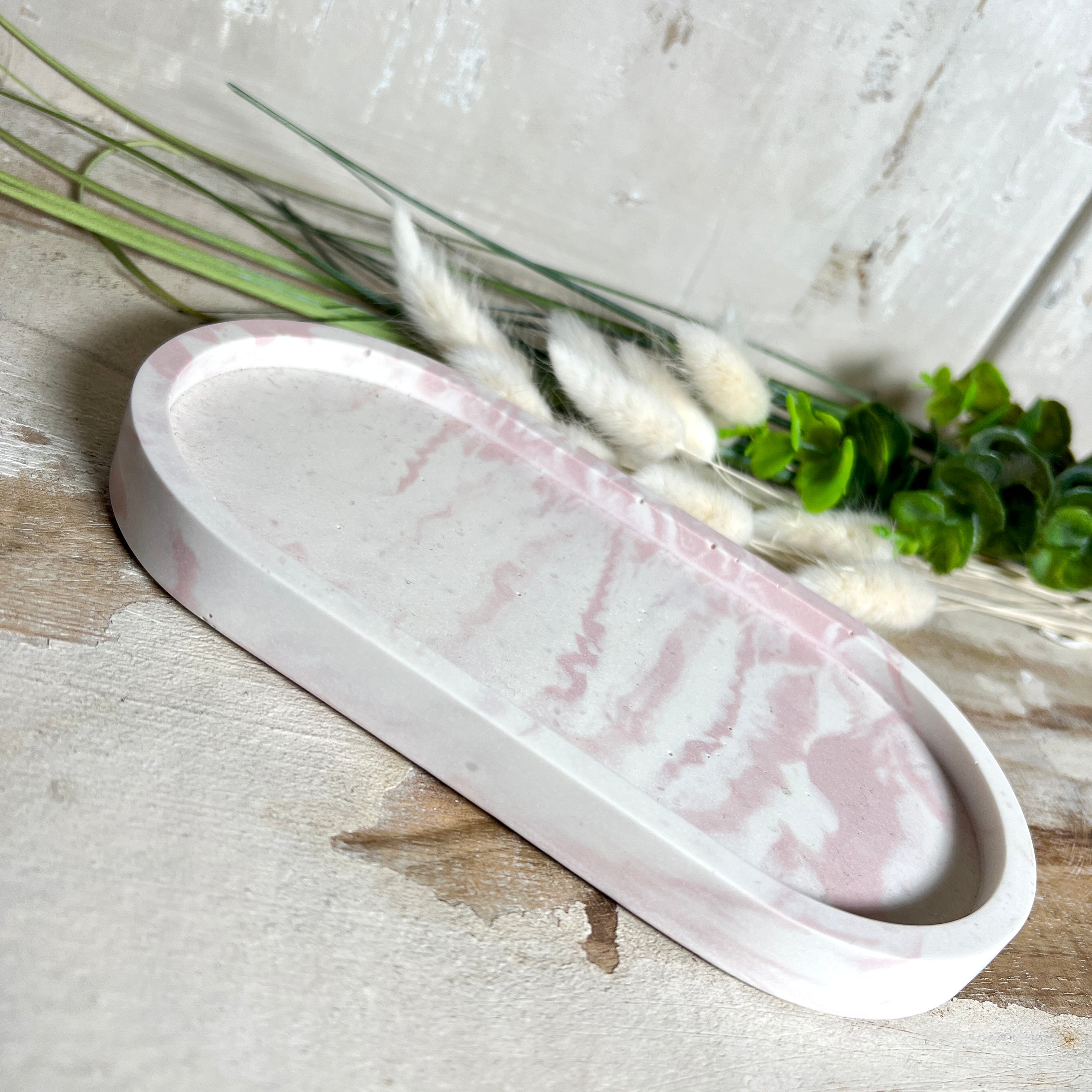 Schmucktablett aus Jesmonite in zartem Rosa-Weiß-Marmor. Handgegossenes, ovales Deko-Tablett mit glatter Oberfläche und Korkfüßen. Perfekt als Ablage für Schmuck, Schlüssel, Brille oder Dekoobjekte. Jedes Stück ein liebevoll gefertigtes Unikat in der Größe 17,5 × 9,5 × 1,7 cm.