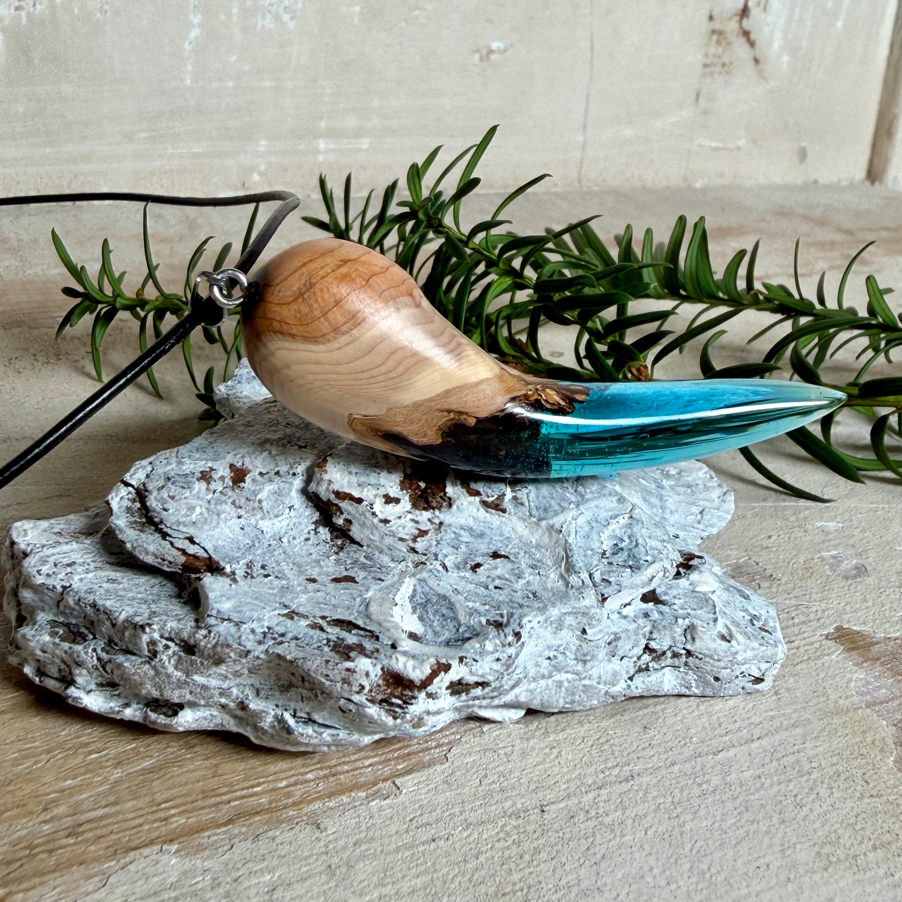 Handgefertigter Anhänger aus Edelholz und transparent-türkisblauem Epoxidharz, glatt poliert. Größe ca. 6,7 × 2,0 cm. Mit Edelstahlöse an schwarzer Lederkette (45 cm + 5 cm Edelstahl-Verlängerung, Karabiner). Maritimer, moderner Unisex-Schmuck mit natürlicher Holzmaserung; kleine Lufteinschlüsse sind charakteristisch.