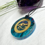 Runder Anhänger mit goldenem Horusauge aus Metall in blau türkis violettem Epoxidharz am schwarzen Baumwollband, spiritueller Eye of Ra Naturschmuck Amulett