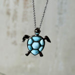 Feine Kette aus rhodiniertem 925 Silber mit Wasserschildkröten-Anhänger. Der ca. 25 x 18 mm große Anhänger zeigt eine Schildkröte, deren Panzer aus mehreren hellblauen Larimar-Inlays besteht. Seltener, kostbarer Larimar mit typischen wolkigen Strukturen, an einer ca. 45 cm langen, 1 mm starken, auf Platin rhodinierten Silberkette. Fotografiert in schwarzer Schmuckbox und vor maritimer Dekoration mit Muscheln.