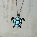 Feine Kette aus rhodiniertem 925 Silber mit Wasserschildkröten-Anhänger. Der ca. 25 x 18 mm große Anhänger zeigt eine Schildkröte, deren Panzer aus mehreren hellblauen Larimar-Inlays besteht. Seltener, kostbarer Larimar mit typischen wolkigen Strukturen, an einer ca. 45 cm langen, 1 mm starken, auf Platin rhodinierten Silberkette. Fotografiert in schwarzer Schmuckbox und vor maritimer Dekoration mit Muscheln.