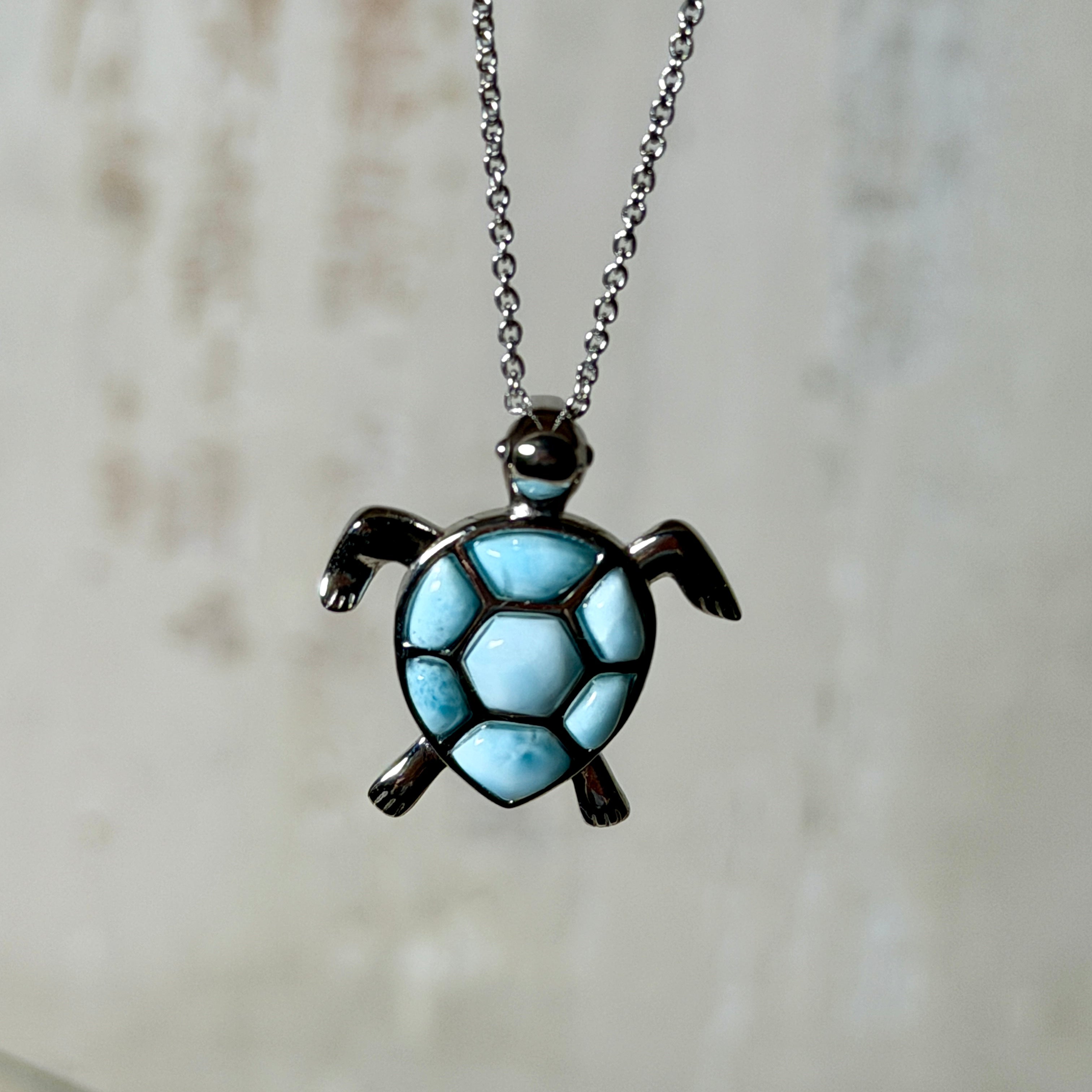 Feine Kette aus rhodiniertem 925 Silber mit Wasserschildkröten-Anhänger. Der ca. 25 x 18 mm große Anhänger zeigt eine Schildkröte, deren Panzer aus mehreren hellblauen Larimar-Inlays besteht. Seltener, kostbarer Larimar mit typischen wolkigen Strukturen, an einer ca. 45 cm langen, 1 mm starken, auf Platin rhodinierten Silberkette. Fotografiert in schwarzer Schmuckbox und vor maritimer Dekoration mit Muscheln.