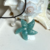 Seestern-Anhänger aus transparentem Harz mit echtem Meersand ( leicht eingefärbt), in blautürkis Strand-Look, mit kleiner Süßwasserperle als Anhänger-Detail. Edelstahlöse, schwarze Lederkette mit Edelstahl-Karabiner und Verlängerung (45 cm + 5 cm). Anhänger ca. 3,5 × 3,5 cm. Handgefertigtes Unikat; vereinzelt kleine Luftbläschen möglich, typisch für Handarbeit.