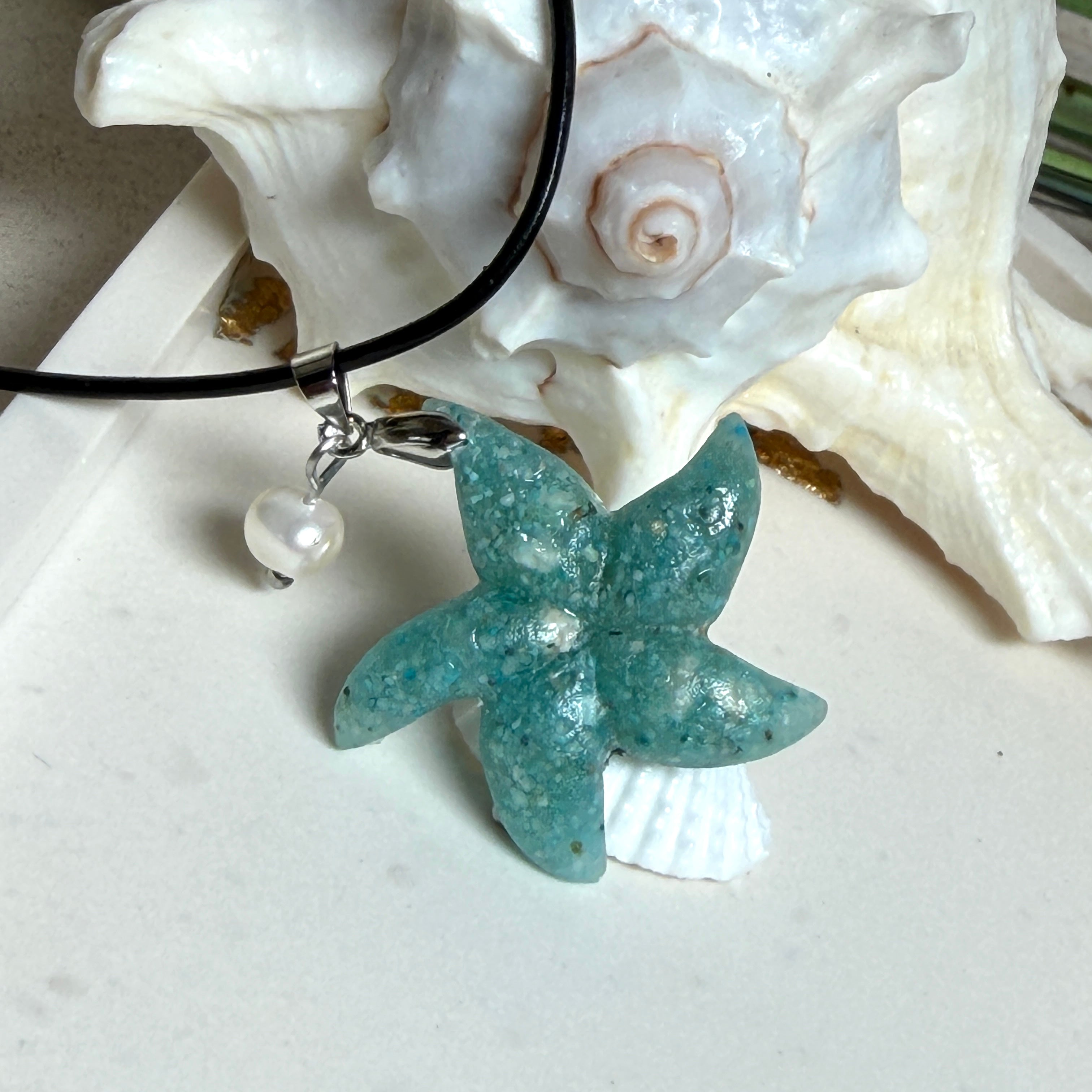 Seestern-Anhänger aus transparentem Harz mit echtem Meersand ( leicht eingefärbt), in blautürkis Strand-Look, mit kleiner Süßwasserperle als Anhänger-Detail. Edelstahlöse, schwarze Lederkette mit Edelstahl-Karabiner und Verlängerung (45 cm + 5 cm). Anhänger ca. 3,5 × 3,5 cm. Handgefertigtes Unikat; vereinzelt kleine Luftbläschen möglich, typisch für Handarbeit.