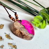 Handgefertigter Herzanhänger aus Olivenholz und leuchtend pinkem Epoxidharz mit eingeschlossenen Pusteblumensamen. Verstellbares braunes Lederband mit Holzperlen, Anhänger ca. 3,5 × 3,3 cm groß. Das Harz leuchtet dezent unter Schwarzlicht. Ein einzigartiges Unikat von ZeitlosSchmuckDesign, inspiriert von Kindheitsglück und Pusteblumen.