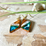 Handgefertigte Ohrringe „Ocean Diamond“ aus Olivenholz und Epoxidharz in Diamantform. Türkis-, Blau- und Weißtöne bilden ein wellenartiges Design, inspiriert vom Meer. Jeder Ohrring ist ein Unikat mit individueller Maserung. Vergoldete, nickelfreie und bleifreie Anhängerschlaufe, 925 Sterling Silber Ohrhaken. Maritimer Schmuck für Meerliebhaber und besondere Anlässe.