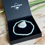 Zartes Larimar-Armband „Lagunenlicht“ mit facettierten 3 mm Perlen aus seltenem Atlantisstein auf Elastikfaden. Die kleinen Larimarsteine zeigen sanfte Blau- und Weißtöne wie Lagunenwasser. Innenumfang ca. 19 cm. Fotografiert auf Muscheln, einem dekorativen Stein und in einer schwarzen Schmuckbox von ZeitlosSchmuckDesign im maritimen Stil.
