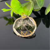 “Handgefertigter Anhänger mit filigranen Pusteblumen-Samen, eingefasst in goldfarbenem Rahmen und präsentiert auf einem glänzenden schwarzen Hintergrund mit grünem Blattdetail. Einzigartiger Schmuck für Naturliebhaber und elegante Styles.”