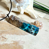 Rechteckiger Stabanhänger aus Maserholz und glasklarem Epoxidharz in tiefen Blau- und Türkistönen mit organischer Lagunen/Riff-Optik. Hochglanzpoliert, ca. 5,3 × 1,2 cm. Der Anhänger hängt an einer schwarzen Lederkette mit Edelstahl-Karabiner und 5-cm-Verlängerung (45 + 5 cm); die Anhängerschlaufe ist ebenfalls aus Edelstahl. Handgefertigtes Unikat, Lieferung in edler Schmuckbox – maritimer Look.