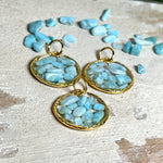 Runder Anhänger „Golden Atlantis Dream“ mit kleinen Larimar-Edelsteinchen in klarem Resin, gefasst in einer 24K vergoldeten Zinklegierung in Shiny Gold. Die Larimarsteine zeigen sanfte Blau- und Weißtöne wie karibische Wellen. Durchmesser ca. 22 mm, handgefertigtes Unikat, fotografiert mit Larimarsteinchen, Muscheln und einer 2-Euro-Münze zur Größenorientierung auf maritimem Untergrund.