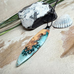 Handgefertigter, länglicher Surfbrett-Anhänger aus naturbelassenem Maserholz und transparentem Harz in Lagunen-Türkis, an schwarzem Lederband mit Knoten – maritimer Unikatschmuck für Damen und Herren, dekorativ mit Stein und Muschel inszeniert.
