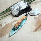 Handgefertigter, länglicher Surfbrett-Anhänger aus naturbelassenem Maserholz und transparentem Harz in Lagunen-Türkis, an schwarzem Lederband mit Knoten – maritimer Unikatschmuck für Damen und Herren, dekorativ mit Stein und Muschel inszeniert.