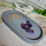 Handgefertigtes ovales Schmucktablett aus grauen  Jesmonite mit schimmernden Blattgold-Details. Die Unterseite ist mit vier Korkpunkten versehen und schützt empfindliche Oberflächen. Ideal zur stilvollen Aufbewahrung von Schmuck, Schlüsseln, Brille oder kleinen Dekoartikeln. Jedes Tablett ist ein Unikat mit individueller Maserung und goldenen Akzenten – Lieferung ohne Deko. Die 