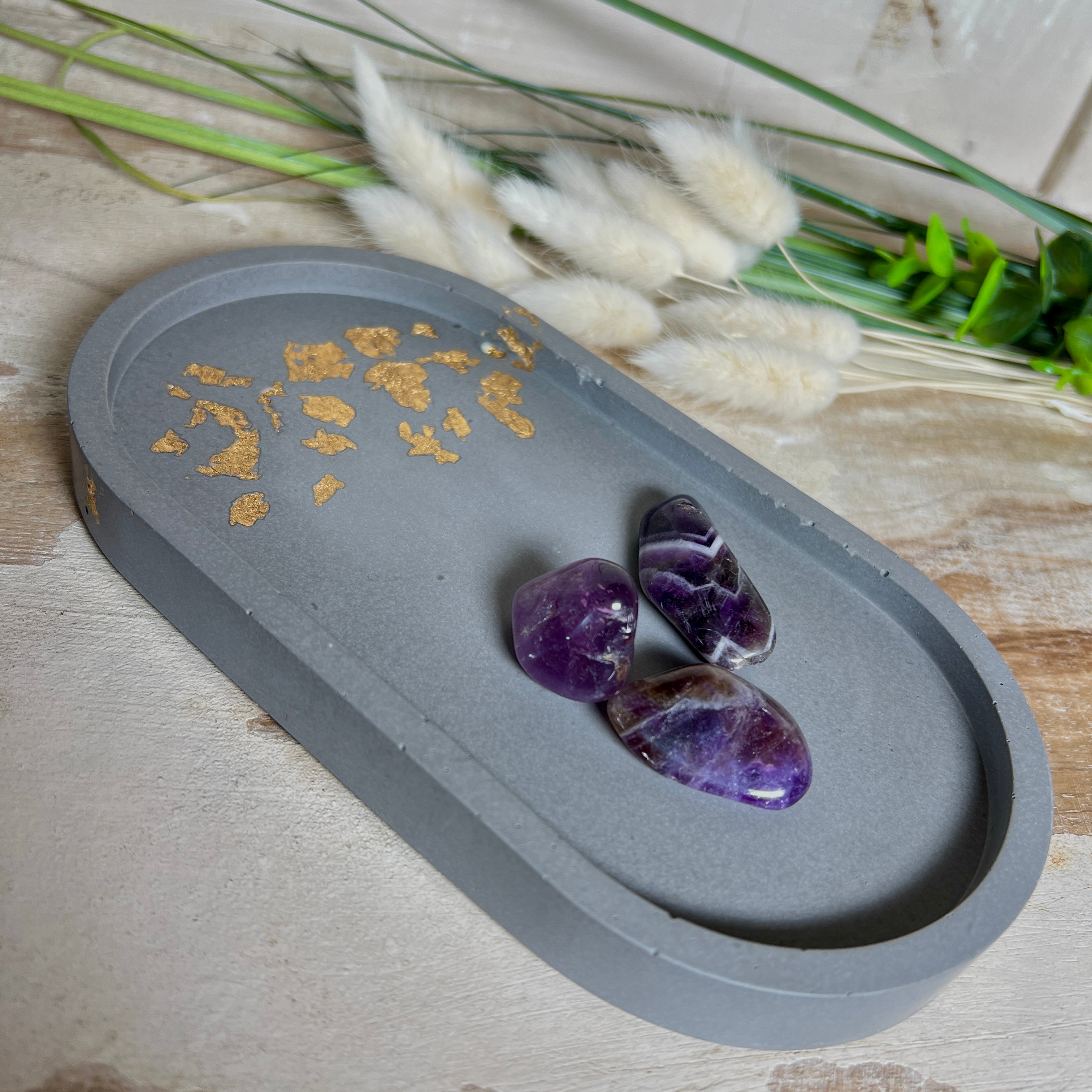 Handgefertigtes ovales Schmucktablett aus grauen  Jesmonite mit schimmernden Blattgold-Details. Die Unterseite ist mit vier Korkpunkten versehen und schützt empfindliche Oberflächen. Ideal zur stilvollen Aufbewahrung von Schmuck, Schlüsseln, Brille oder kleinen Dekoartikeln. Jedes Tablett ist ein Unikat mit individueller Maserung und goldenen Akzenten – Lieferung ohne Deko. Die 