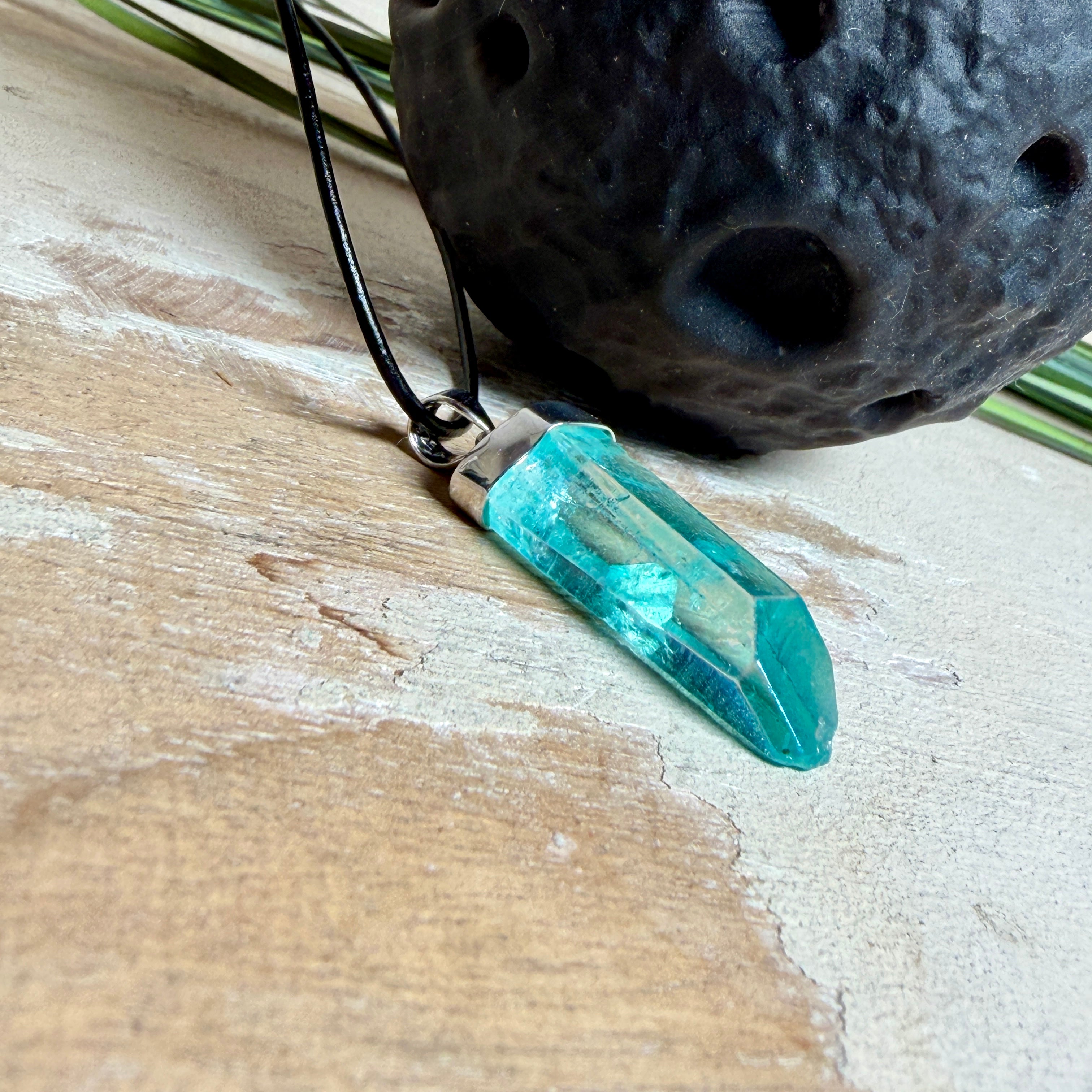 Nahaufnahme einer handgefertigten Halskette mit Aqua Aura Rohkristall: naturbelassener, sechskantiger Bergkristall mit goldbedampfter Aqua-Beschichtung in Türkisblau, gefasst in 925er Silber und getragen an einer schwarzen Lederkette mit Edelstahl-Karabiner und Verlängerungskettchen.