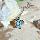 Feine Kette aus rhodiniertem 925 Silber mit Wasserschildkröten-Anhänger. Der ca. 25 x 18 mm große Anhänger zeigt eine Schildkröte, deren Panzer aus mehreren hellblauen Larimar-Inlays besteht. Seltener, kostbarer Larimar mit typischen wolkigen Strukturen, an einer ca. 45 cm langen, 1 mm starken, auf Platin rhodinierten Silberkette. Fotografiert in schwarzer Schmuckbox und vor maritimer Dekoration mit Muscheln.