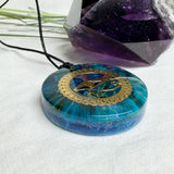 Runder Anhänger mit goldenem Horusauge aus Metall in blau türkis violettem Epoxidharz am schwarzen Baumwollband, spiritueller Eye of Ra Naturschmuck Amulett