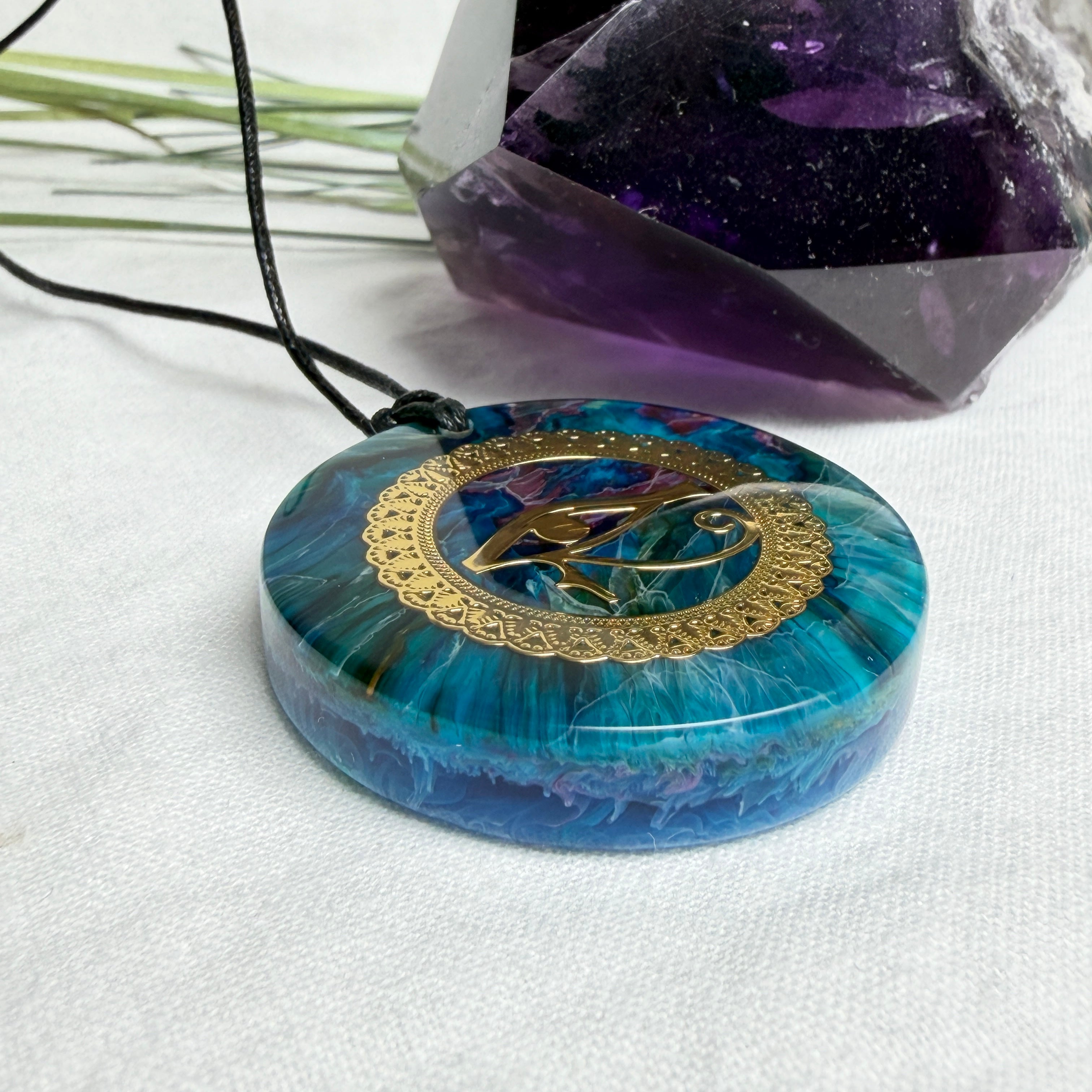 Runder Anhänger mit goldenem Horusauge aus Metall in blau türkis violettem Epoxidharz am schwarzen Baumwollband, spiritueller Eye of Ra Naturschmuck Amulett