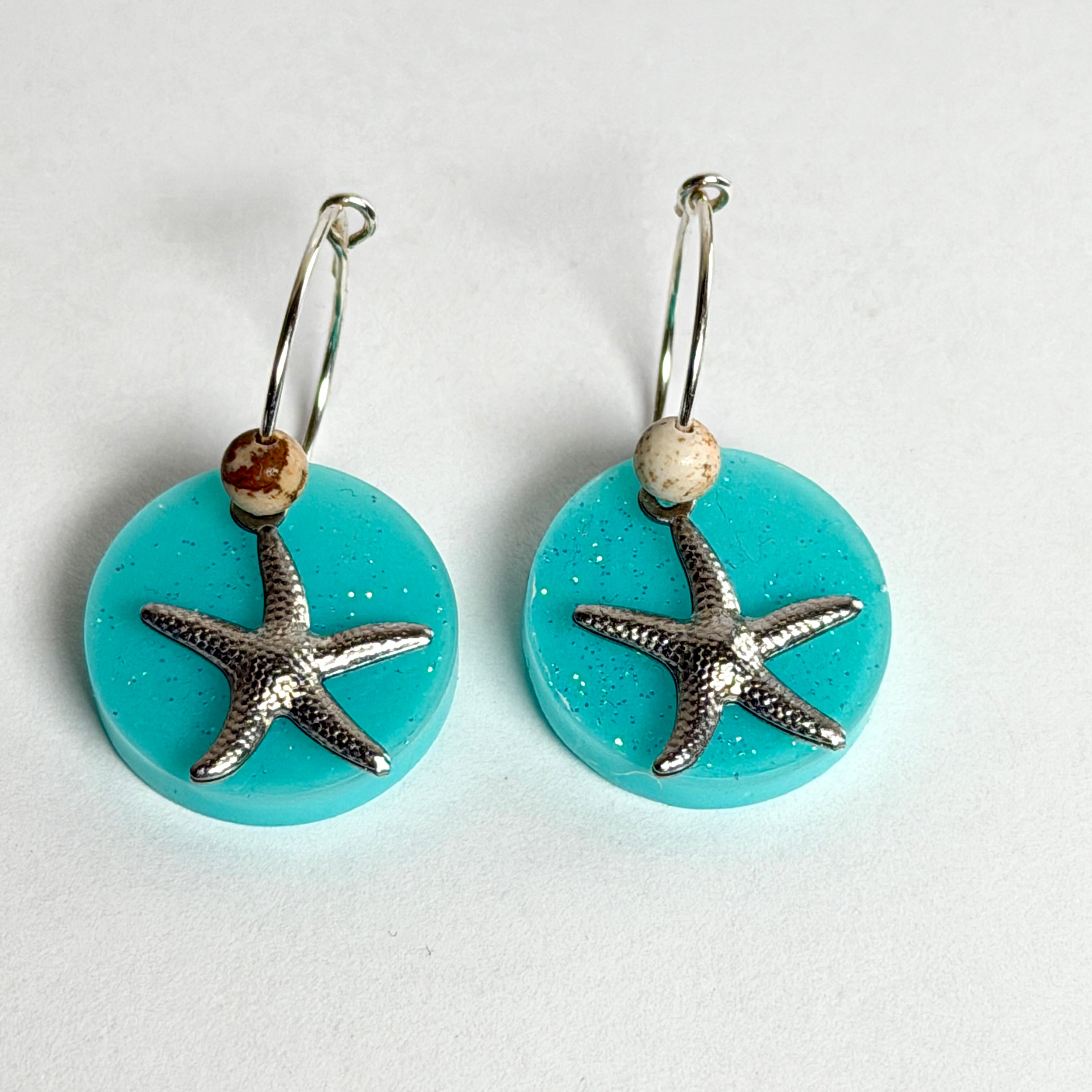 Handgefertigte Creolen-Ohrringe mit türkisblauer Resin-Scheibe, Glitzereffekt, silbernem Seestern-Charm und Jaspis-Natursteinperle. 925 Silber Creole, Gesamtlänge ca. 3,5 cm – Ocean-Feeling für jeden Tag.