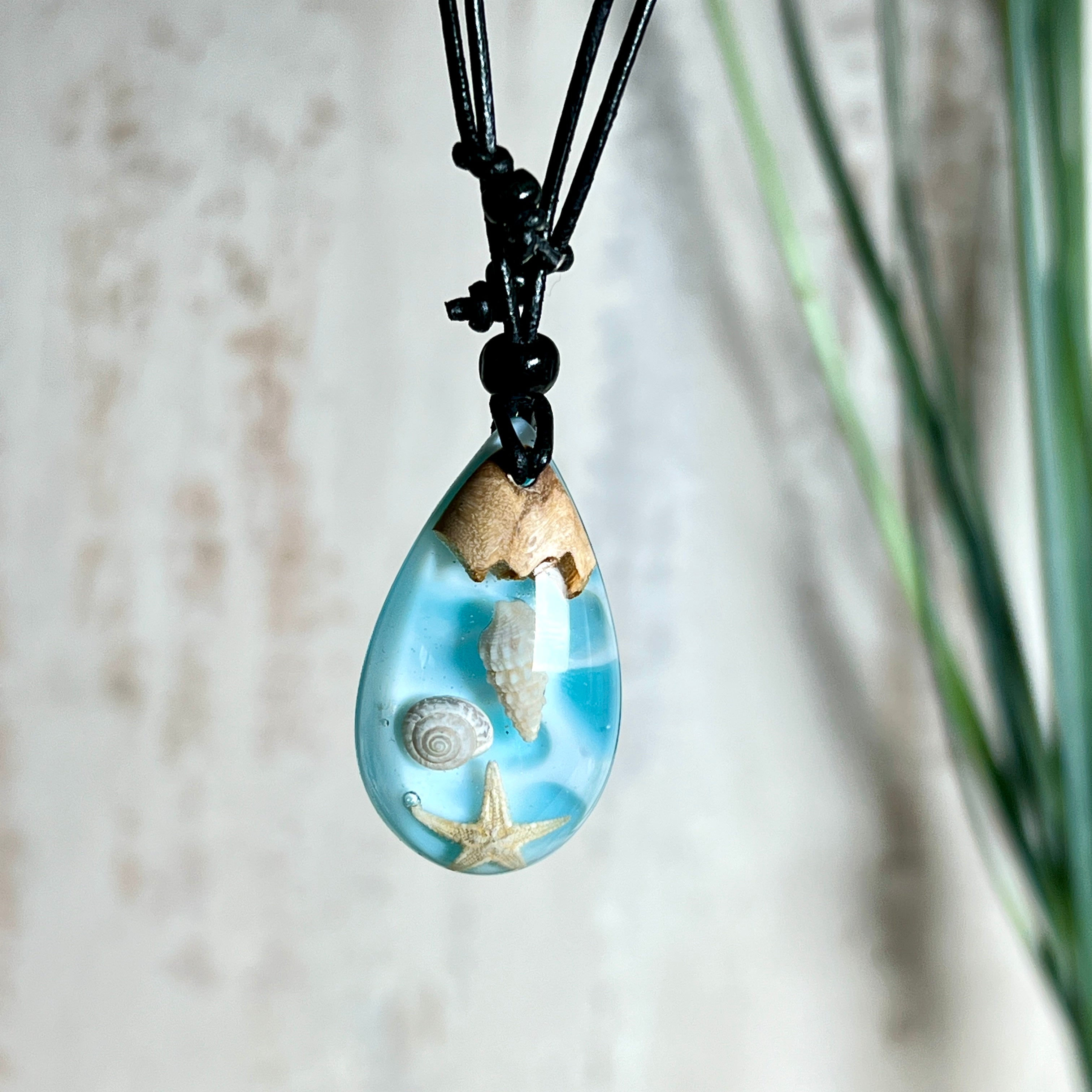 Handgefertigter Tropfenanhänger aus Maserholz und klarem Epoxidharz in Aquatönen. Im Harz sind echte Muscheln und ein kleiner Seestern eingebettet. Durch die besondere Mehrschicht-Gießtechnik wirkt es, als würden die Muscheln im Wasser schweben. Größe ca. 3,8 × 2 cm. Verstellbares Lederband (24–44 cm) mit Holzperlen. Maritimes Unikat.