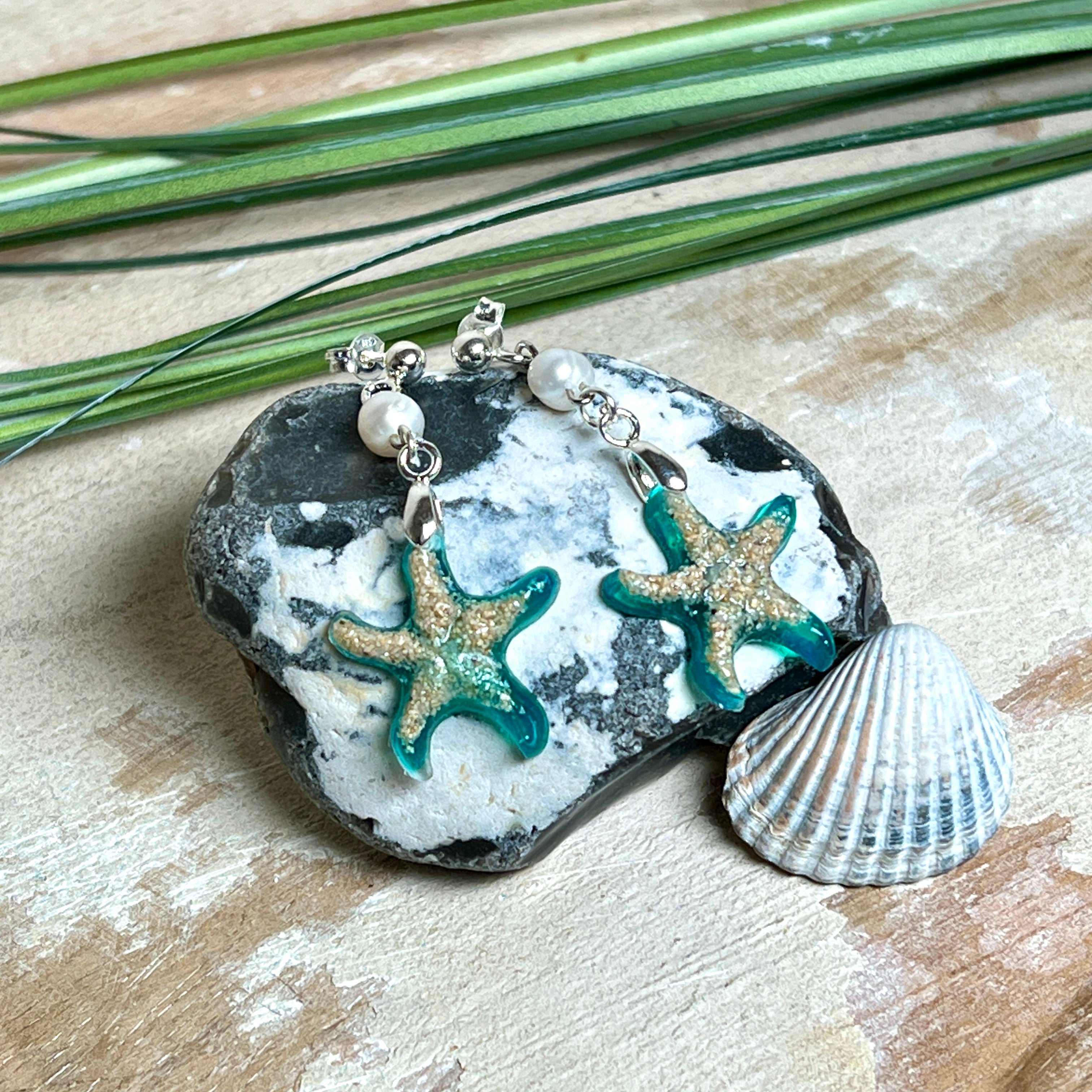 Handgefertigte Seestern-Ohrringe aus Epoxidharz und Sand mit Süßwasserperle, an 925 Sterling Silber Ohrsteckern; versilberte Anhängerschlaufe. Je Seestern ca. 2,6 × 2,5 cm. Leichte, maritime Dangle-Ohrringe im Strand-Look – ideal als Geschenk für Meeresliebhaberinnen.