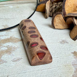 Handgefertigter Halsanhänger aus echtem Banksiazapfen, veredelt mit rotem Harz in den natürlichen Kammern; ca. 5,5 × 1,6 cm. Mit Anhängerschlaufe aus Edelstahl an schwarzer Lederkette (45 cm + 5 cm Verlängerung) mit Edelstahl-Karabiner. Detailaufnahme bzw. Tragebild vor neutralem, natürlichem Hintergrund.