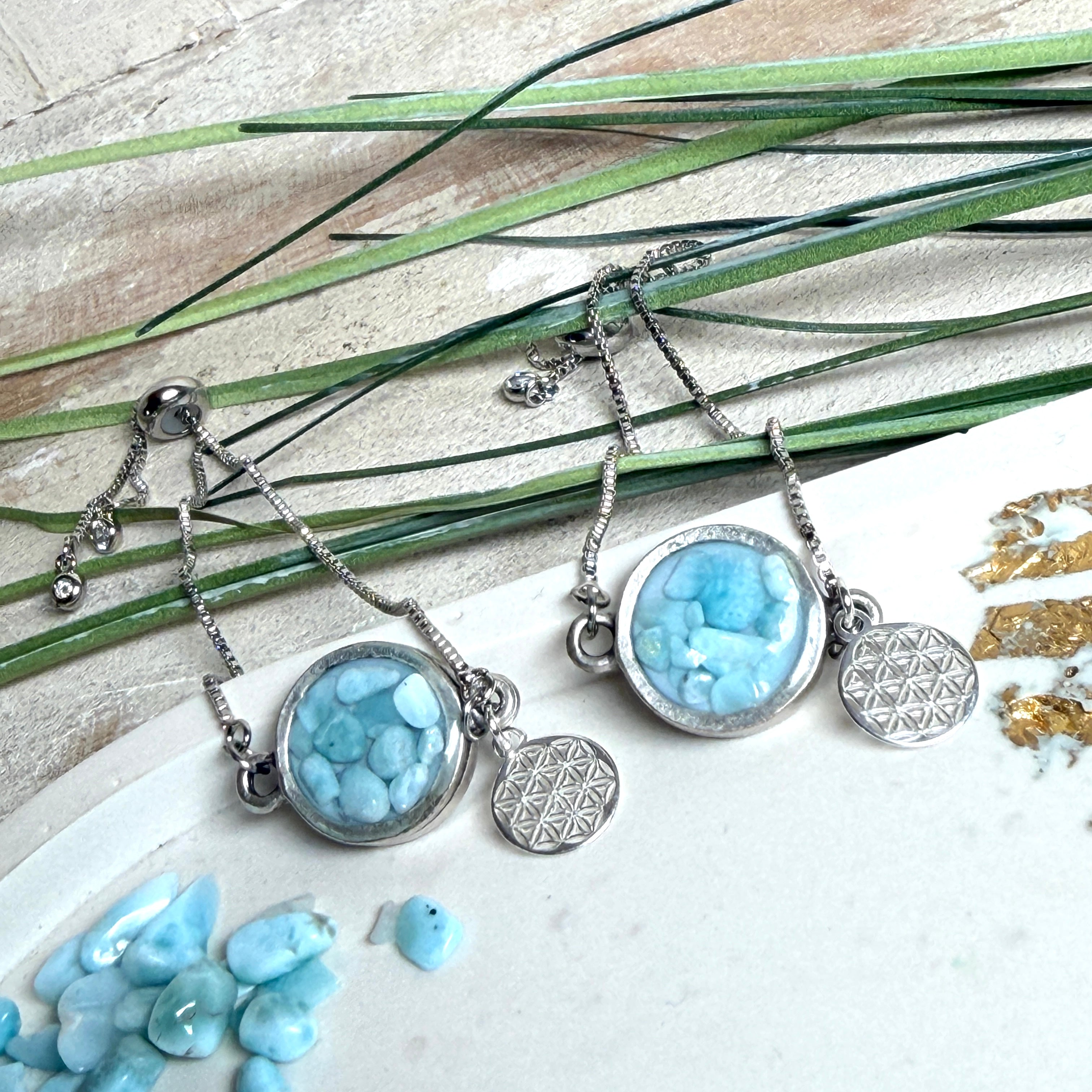 Verstellbares Edelstahlarmband mit Zirkoniasteinchen und Schiebeverschluss und rundem Anhänger (ca. 2,5 × 1,7 cm) mit echten Larimar-Edelstein-Splittern in Harz. Zusätzlich ein Charm „Blume des Lebens“ aus 925 Silber. Handgefertigtes Unikat in Türkis- und Meeresblautönen – Schmuckstück in Nahaufnahme bzw. als Tragebild am Handgelenk.