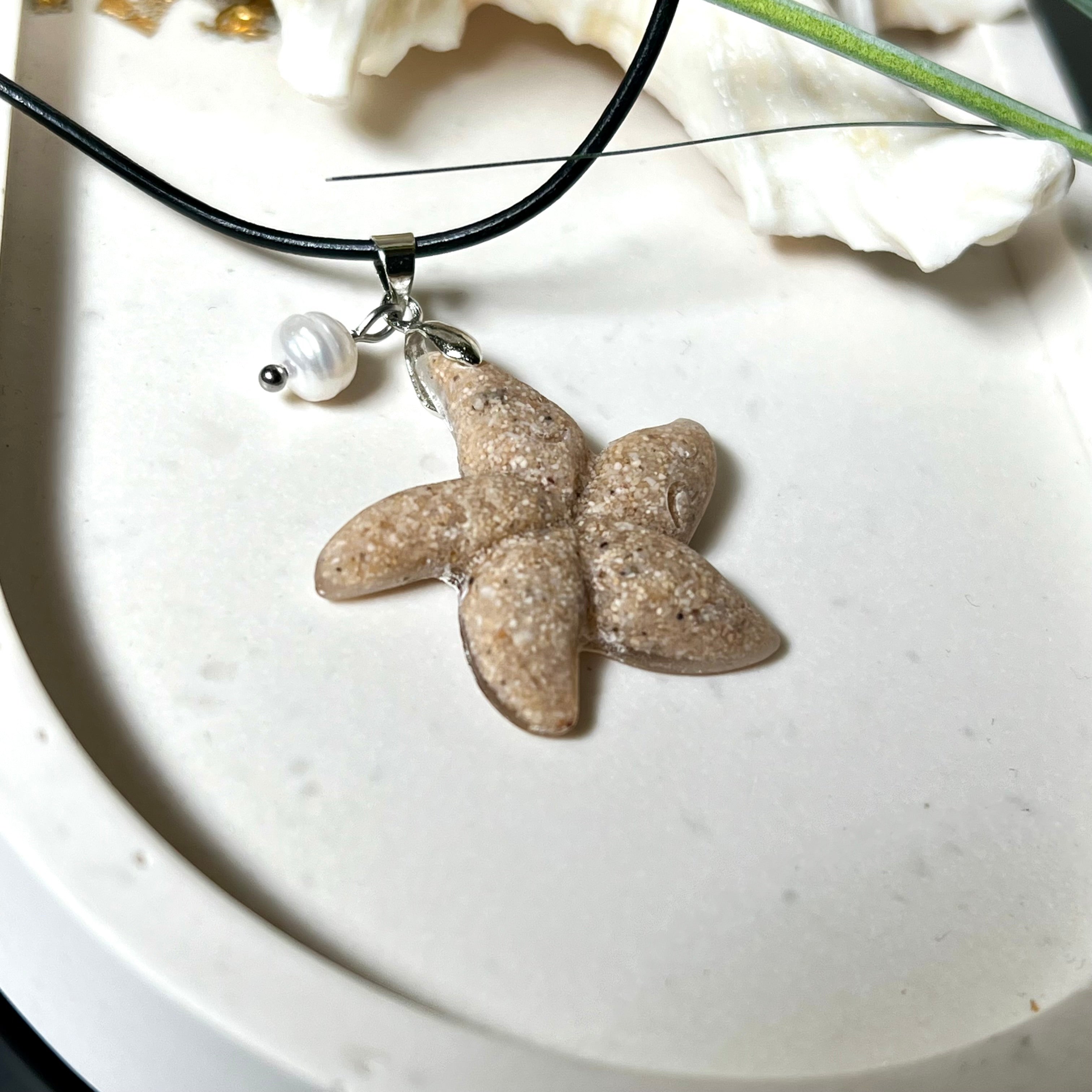 Handgefertigter Anhänger in Seesternform aus echtem Meersand und transparentem Harz. Verziert mit einer natürlichen Süßwasserperle. Edelstahlöse, Lederkette mit Karabinerverschluss (45 cm + 5 cm Verlängerung). Maritimer Schmuck im Unikatcharakter – inspiriert vom Meer und handgefertigt mit Liebe zum Detail. Perfektes Geschenk für Strandliebhaber.