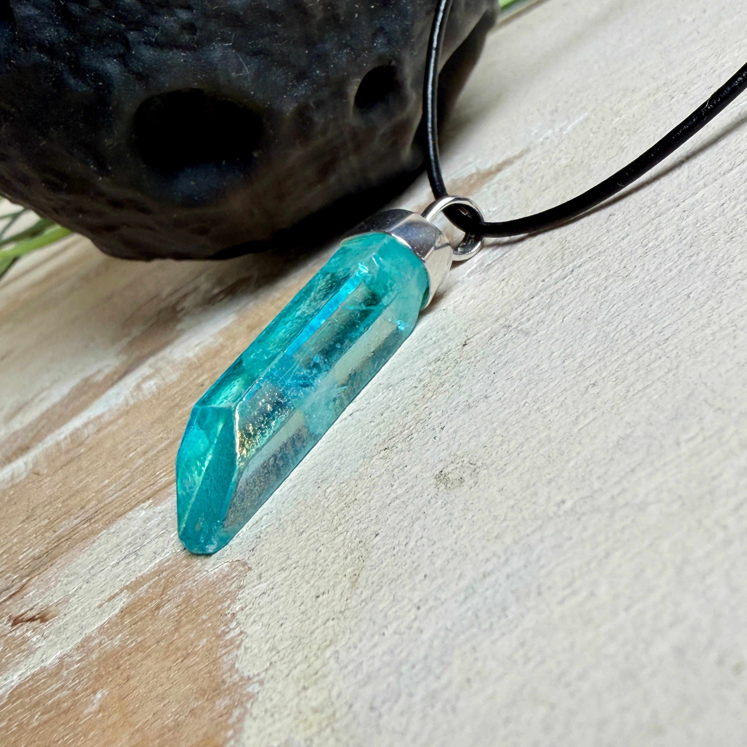 Nahaufnahme einer handgefertigten Halskette mit Aqua Aura Rohkristall: naturbelassener, sechskantiger Bergkristall mit goldbedampfter Aqua-Beschichtung in Türkisblau, gefasst in 925er Silber und getragen an einer schwarzen Lederkette mit Edelstahl-Karabiner und Verlängerungskettchen.