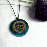 Runder Anhänger mit goldenem Horusauge aus Metall in blau türkis violettem Epoxidharz am schwarzen Baumwollband, spiritueller Eye of Ra Naturschmuck Amulett
