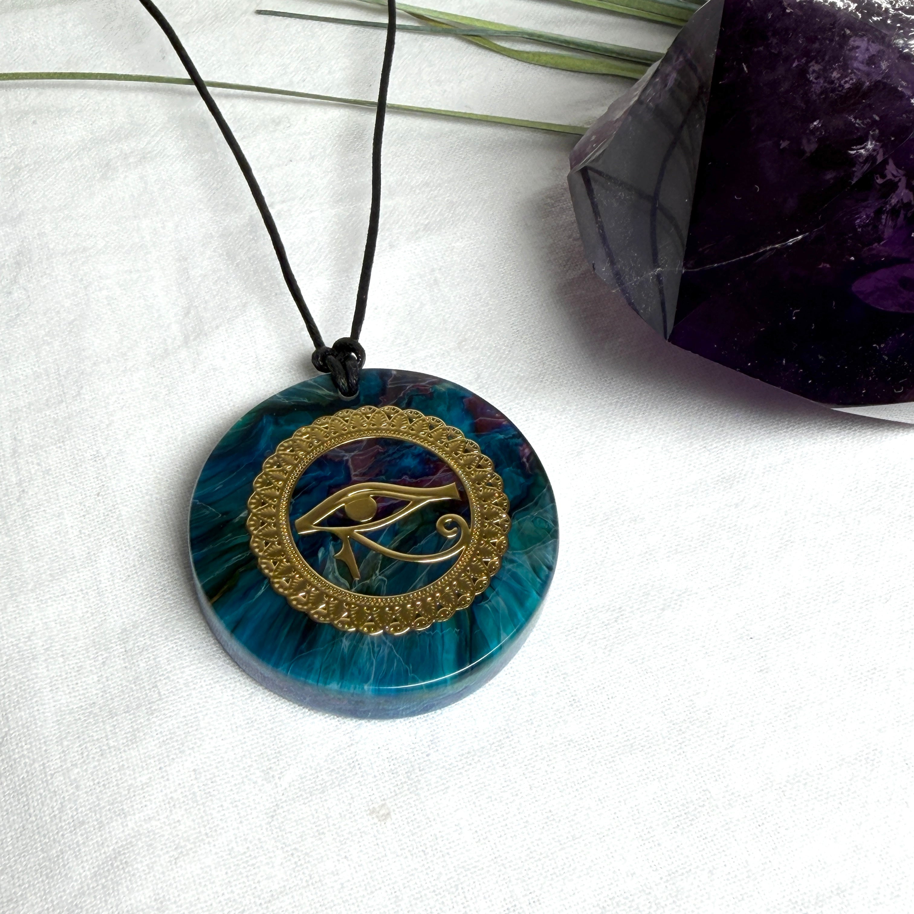 Runder Anhänger mit goldenem Horusauge aus Metall in blau türkis violettem Epoxidharz am schwarzen Baumwollband, spiritueller Eye of Ra Naturschmuck Amulett