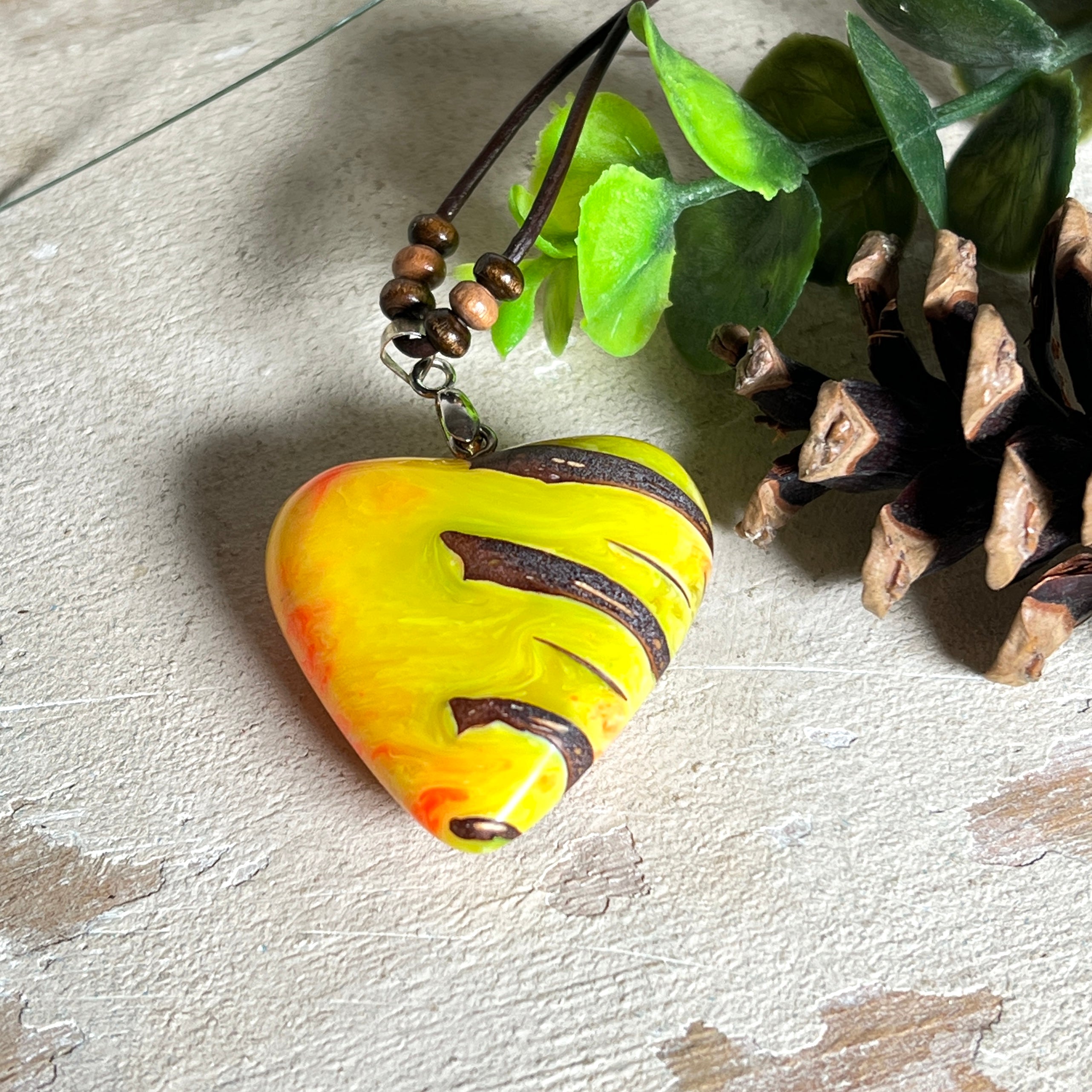 Herz-Anhänger aus Pinienzapfen & Harz, fluoreszierend bei Schwarzlicht. Farben Gelb-Orange-Weiß, Größe 4,2 × 3,8 cm. Lederband verstellbar.