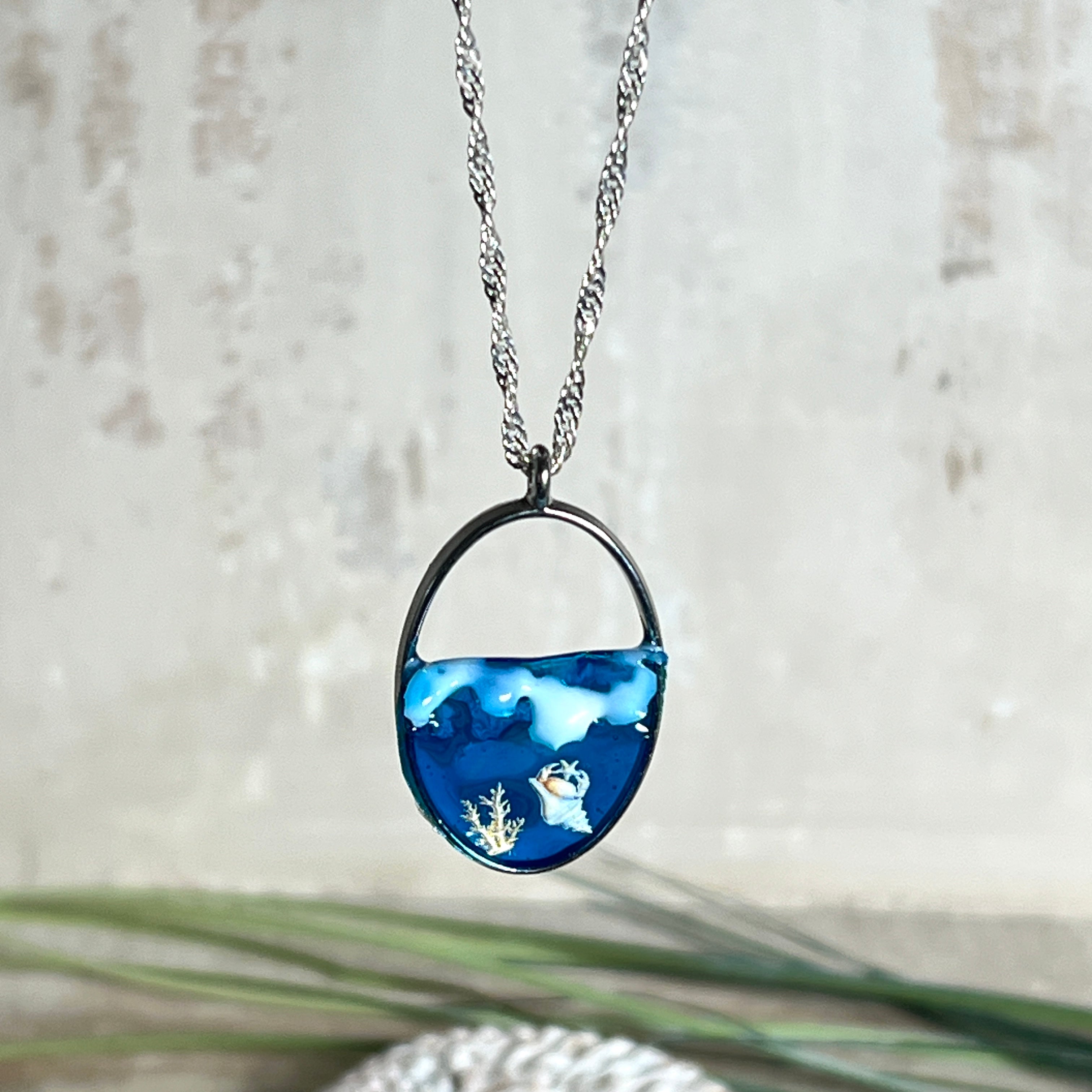 „Riffschatz“ – ovaler Harz-Anhänger mit plastischer 3D-Welle in Blau/Türkis, weißer Schaumkrone und Muschel-/Korallen-Stickern am Grund. Die Sticker sind unter klarem Harz versiegelt. Silber-Legierung, ca. 3,9 × 2,3 cm, an einer ca. 56 cm langen versilberten Kette. Handgefertigt, hochglänzend, jedes Stück ein Unikat.