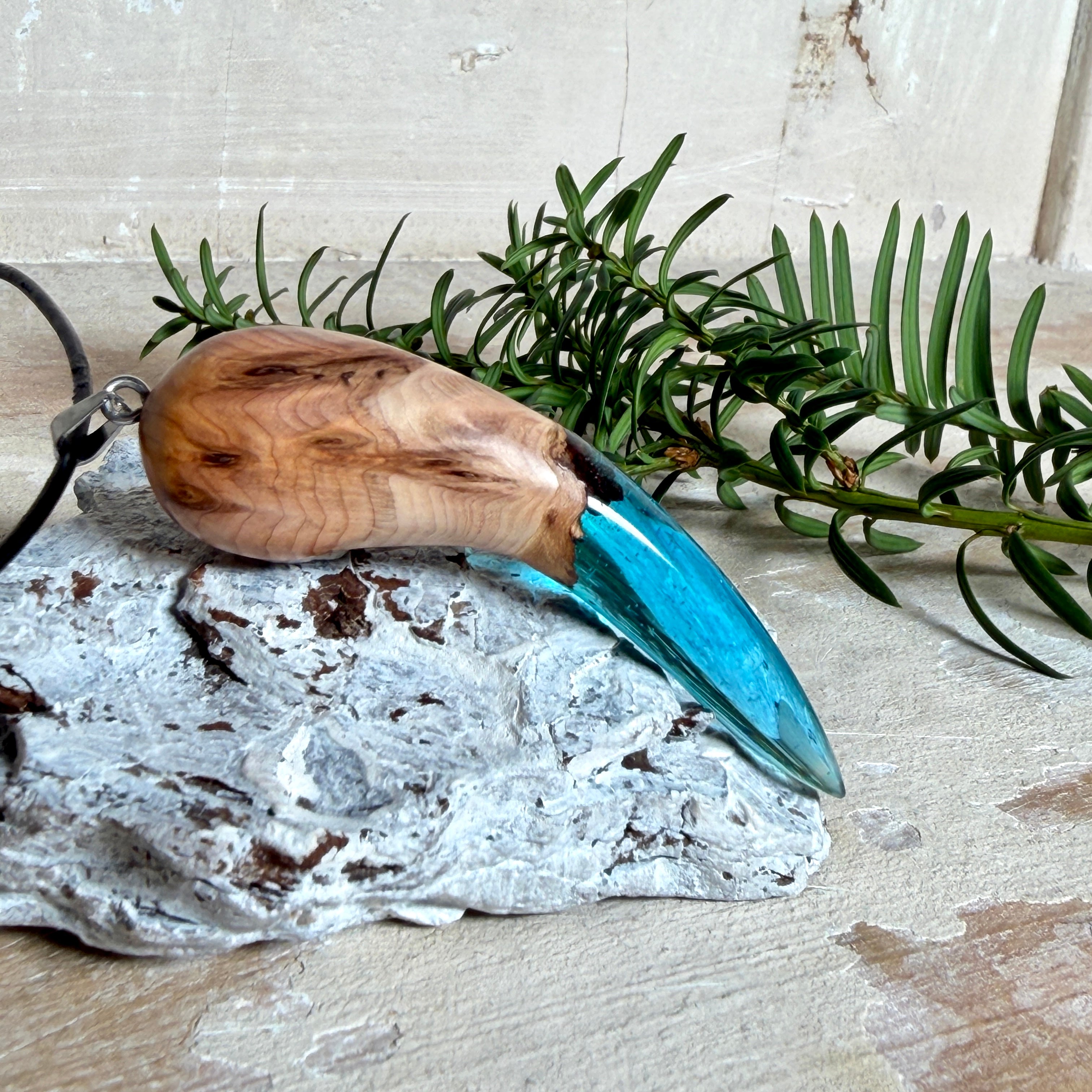 Handgefertigter Anhänger aus Edelholz und transparent-türkisblauem Epoxidharz, glatt poliert. Größe ca. 6,7 × 2,0 cm. Mit Edelstahlöse an schwarzer Lederkette (45 cm + 5 cm Edelstahl-Verlängerung, Karabiner). Maritimer, moderner Unisex-Schmuck mit natürlicher Holzmaserung; kleine Lufteinschlüsse sind charakteristisch.