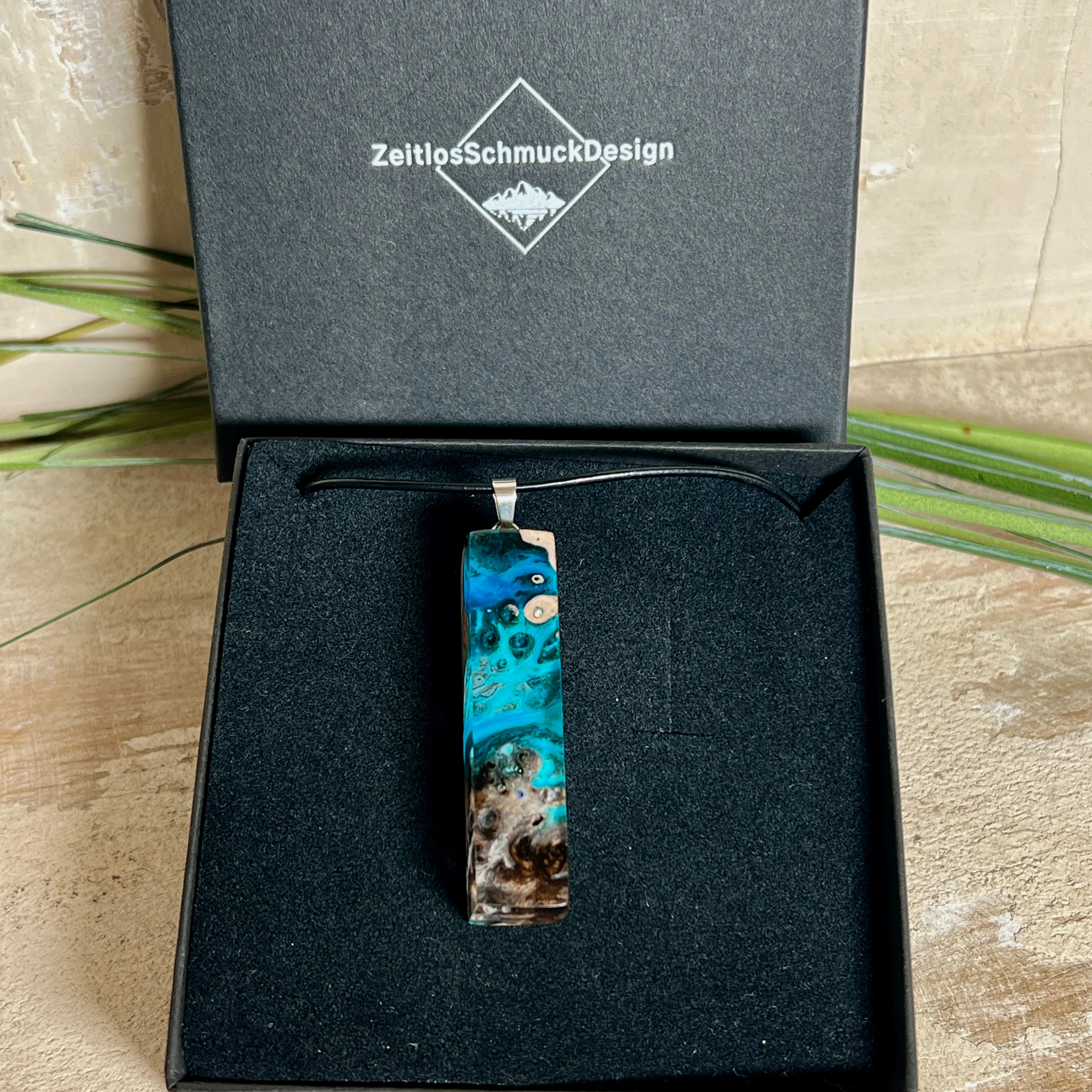 Rechteckiger Stabanhänger aus Maserholz und glasklarem Epoxidharz in tiefen Blau- und Türkistönen mit organischer Lagunen/Riff-Optik. Hochglanzpoliert, ca. 4,6 × 1,2 cm. Der Anhänger hängt an einer schwarzen Lederkette mit Edelstahl-Karabiner und 5-cm-Verlängerung (45 + 5 cm); die Anhängerschlaufe ist ebenfalls aus Edelstahl. Handgefertigtes Unikat, Lieferung in edler Schmuckbox – maritimer Look.
