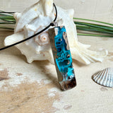 Rechteckiger Stabanhänger aus Maserholz und glasklarem Epoxidharz in tiefen Blau- und Türkistönen mit organischer Lagunen/Riff-Optik. Hochglanzpoliert, ca. 5,3 × 1,2 cm. Der Anhänger hängt an einer schwarzen Lederkette mit Edelstahl-Karabiner und 5-cm-Verlängerung (45 + 5 cm); die Anhängerschlaufe ist ebenfalls aus Edelstahl. Handgefertigtes Unikat, Lieferung in edler Schmuckbox – maritimer Look.