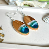 Handgefertigte Ohrringe „Wave Harmony“ aus Olivenholz und Epoxidharz in Blau-, Türkis- und Weißtönen. Das Design erinnert an Meereswellen am Strand. Jeder Ohrring ist ein Unikat mit individueller Maserung. Versilberte, nickelfreie und bleifreie Anhängerschlaufe, 925 Silber Ohrhaken. Maritimer Schmuck – perfekt für Meerliebhaber und als edles Geschenk.