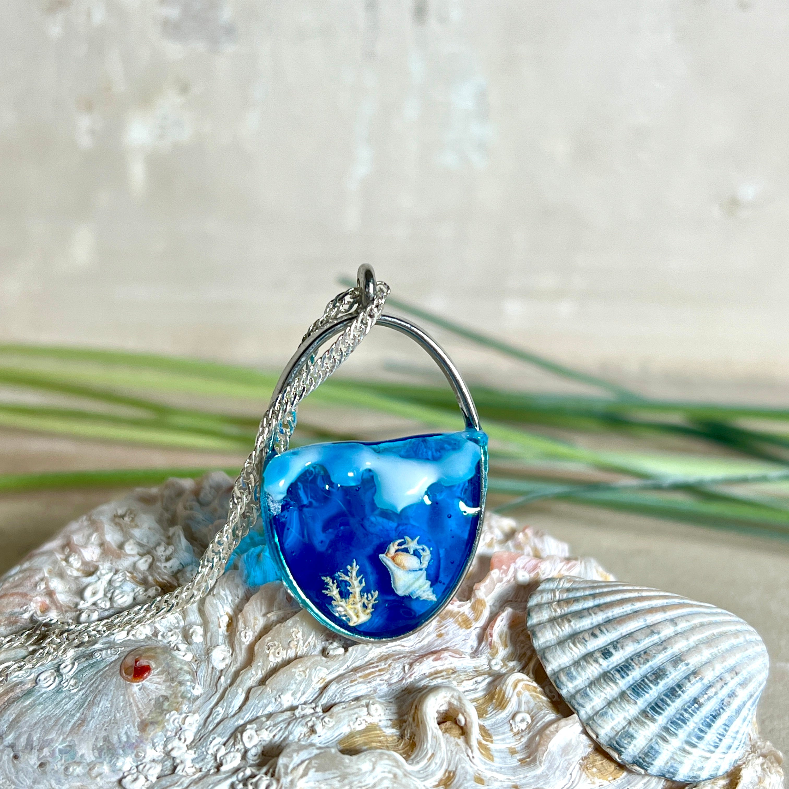 „Riffschatz“ – ovaler Harz-Anhänger mit plastischer 3D-Welle in Blau/Türkis, weißer Schaumkrone und Muschel-/Korallen-Stickern am Grund. Die Sticker sind unter klarem Harz versiegelt. Silber-Legierung, ca. 3,9 × 2,3 cm, an einer ca. 56 cm langen versilberten Kette. Handgefertigt, hochglänzend, jedes Stück ein Unikat.