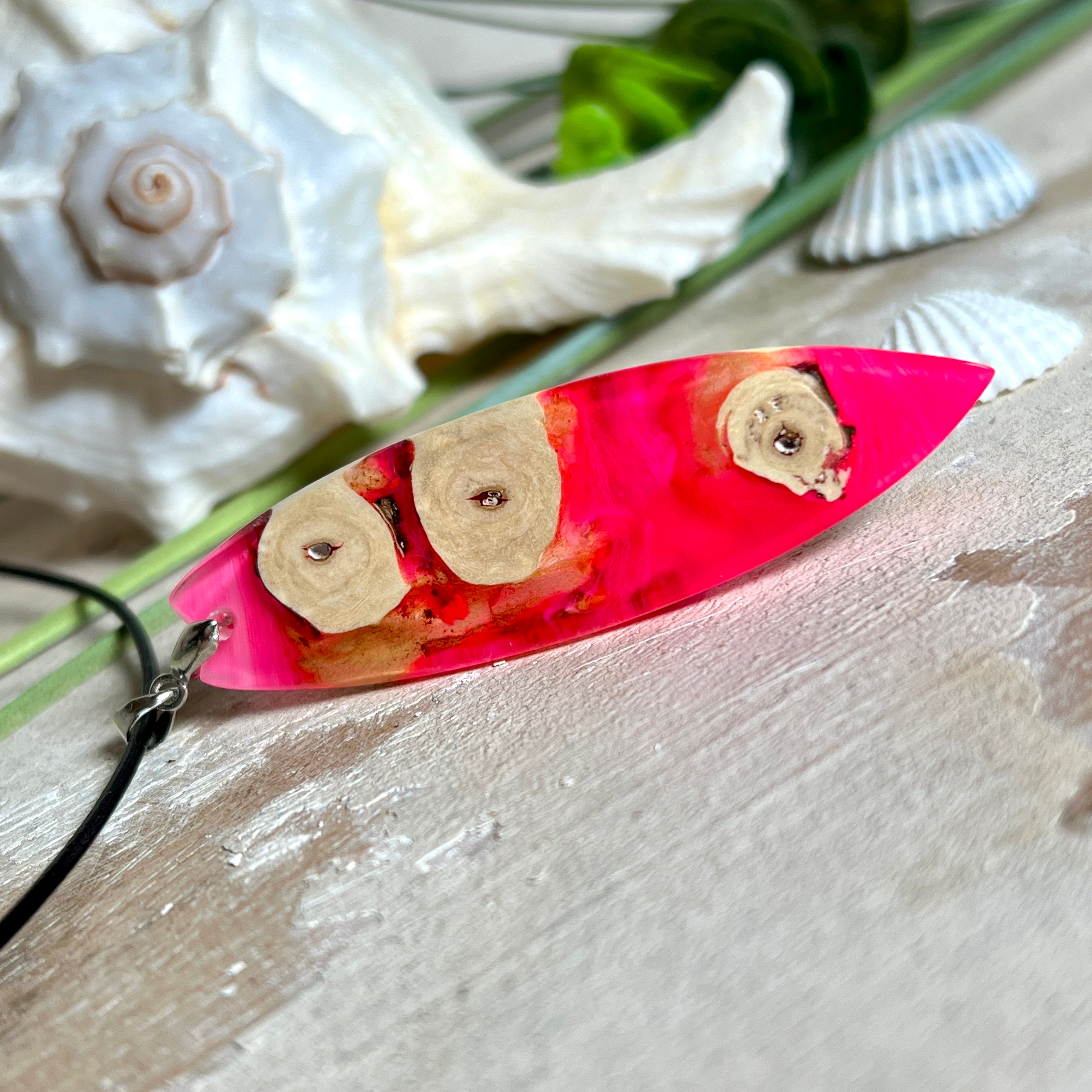 Handgefertigter Surfbrett-Anhänger „Flame Surf“ aus Maserholz und fluoreszierendem Harz in kräftigem Pink. Das Schmuckstück leuchtet bei Schwarzlicht und wird an einer schwarzen Lederkette mit Edelstahl-Karabiner (45 cm + 5 cm Verlängerung) geliefert. Größe ca. 6,7 × 2 cm. Ein Unikat für Surf- und Strandliebhaber, edel verarbeitet und mit Liebe gefertigt.